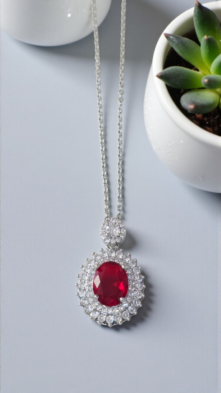 Silver necklace with a red gemstone pendant on a light gray surface with a small plant.