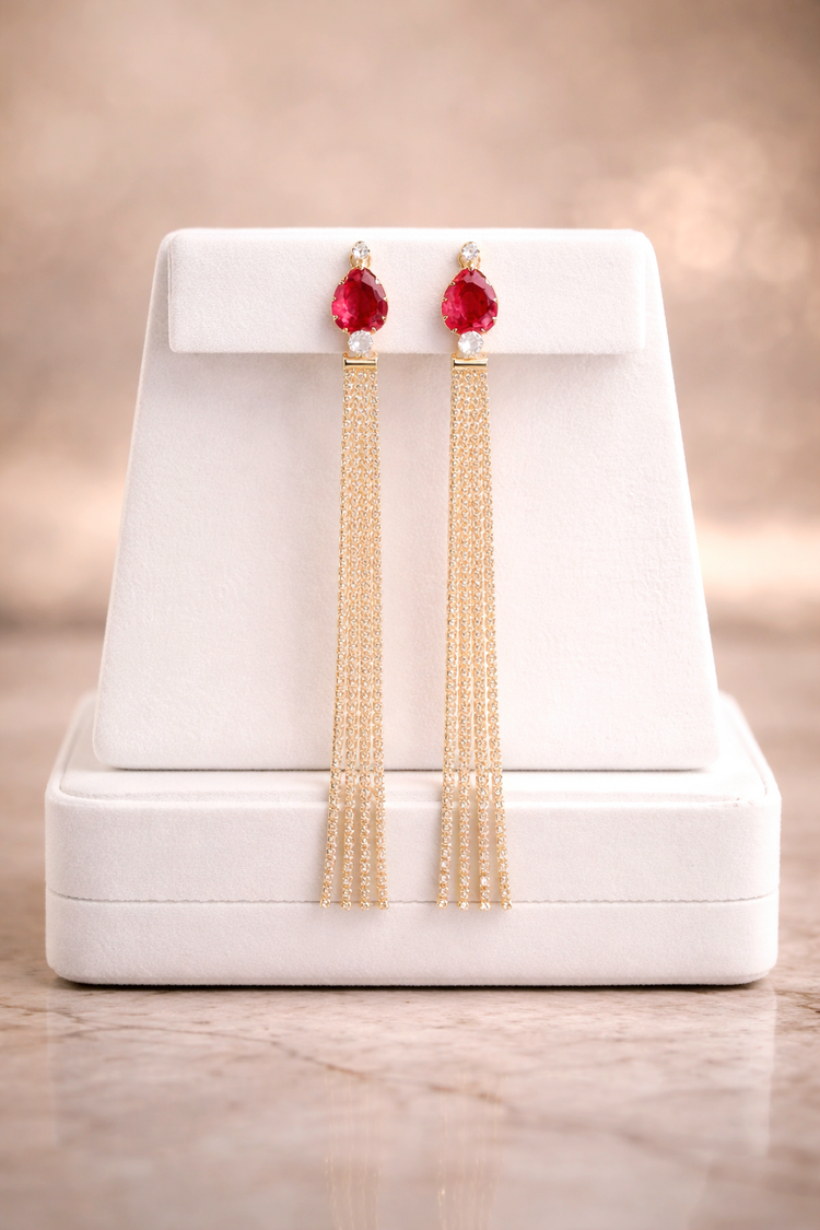 Gold earrings with red stones on a white stand against a beige background