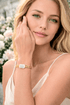 Woman wearing a gold bracelet with a clear stone, outdoors with blurred flowers in the background