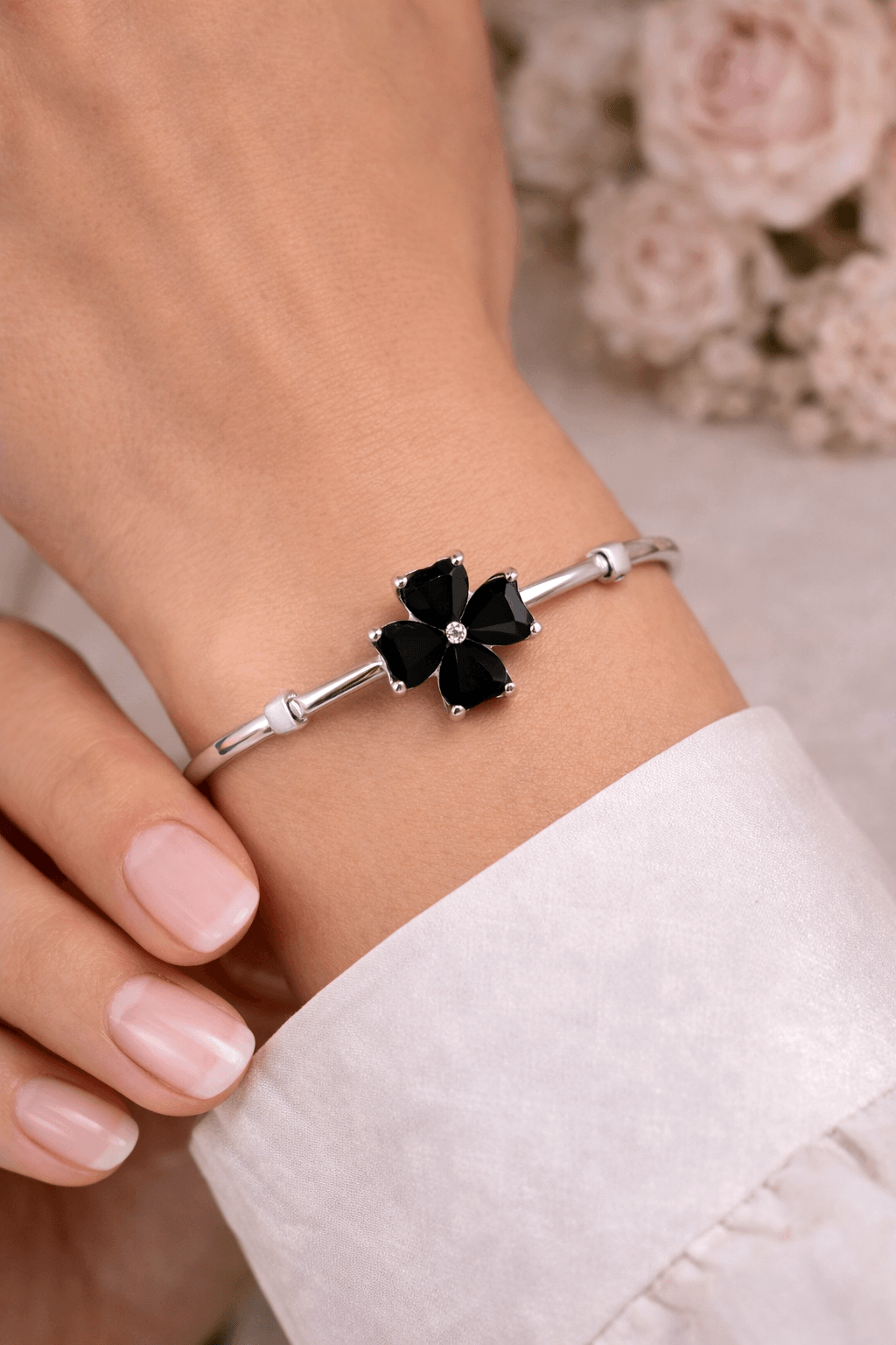Silver bracelet with black clover-shaped charm on a wrist, with a blurred floral background.
