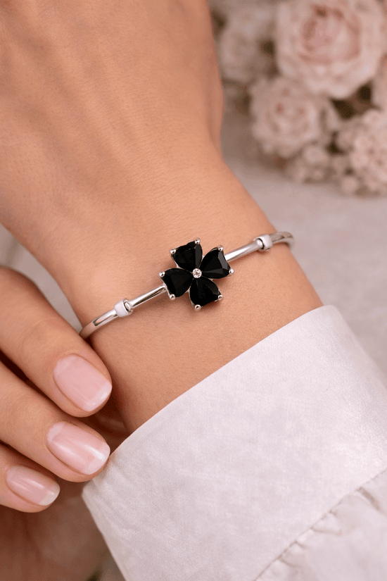 Silver bracelet with black clover-shaped charm on a wrist, with a blurred floral background.