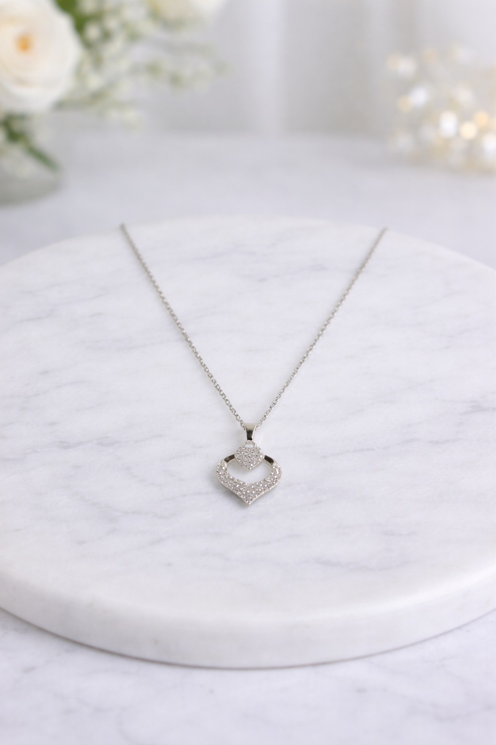 Silver necklace with a heart-shaped pendant on a marble surface with flowers in the background