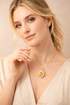 Woman wearing a gold necklace with a sunflower pendant against a beige background