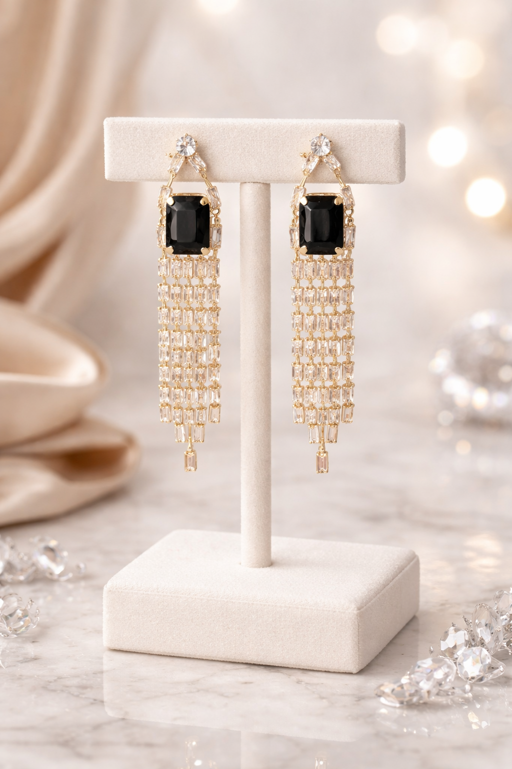 Gold chandelier earrings with black stones on a white stand against a blurred decorative background.