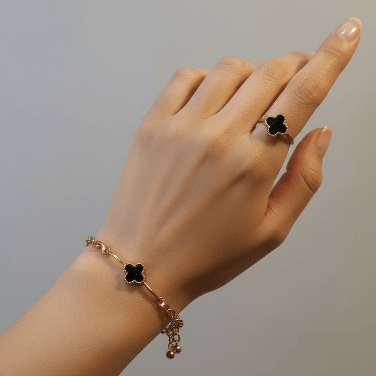 Hand wearing a gold bracelet and ring with black clover designs on a neutral background