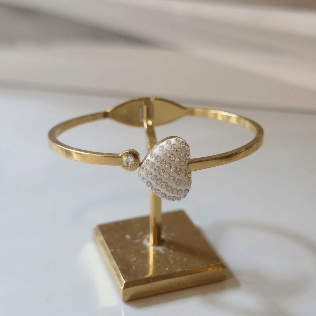 Gold bracelet with a heart-shaped clasp on a matching stand against a light background