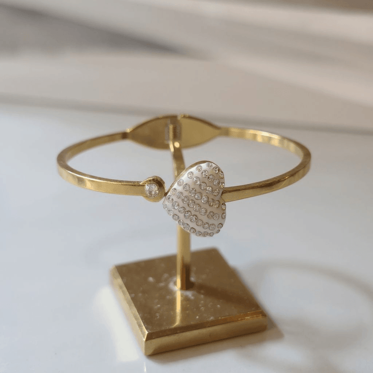 Gold bracelet with a heart-shaped clasp on a matching stand against a light background