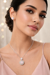 Woman wearing a silver necklace with a pink pendant and matching earrings against a blurred background.