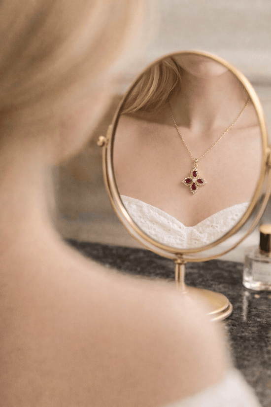 Gold necklace with a red pendant on a small round mirror held by a person.