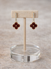 Pair of maroon flower-shaped earrings on a beige stand with a clear circular base.