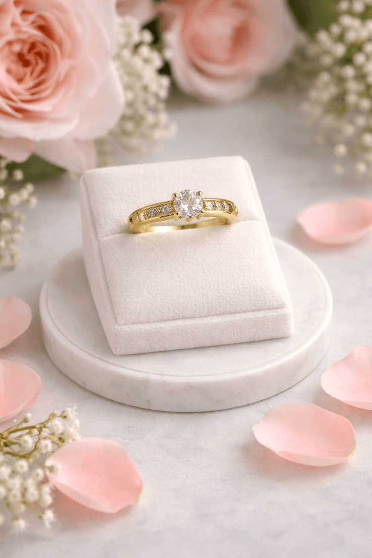 Gold ring in a white box with pink roses and petals on a marble surface