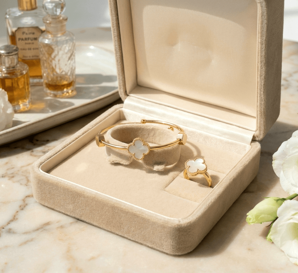 Gold bracelet and ring set in an open jewelry box on a marble surface with flowers and perfume bottles in the background.