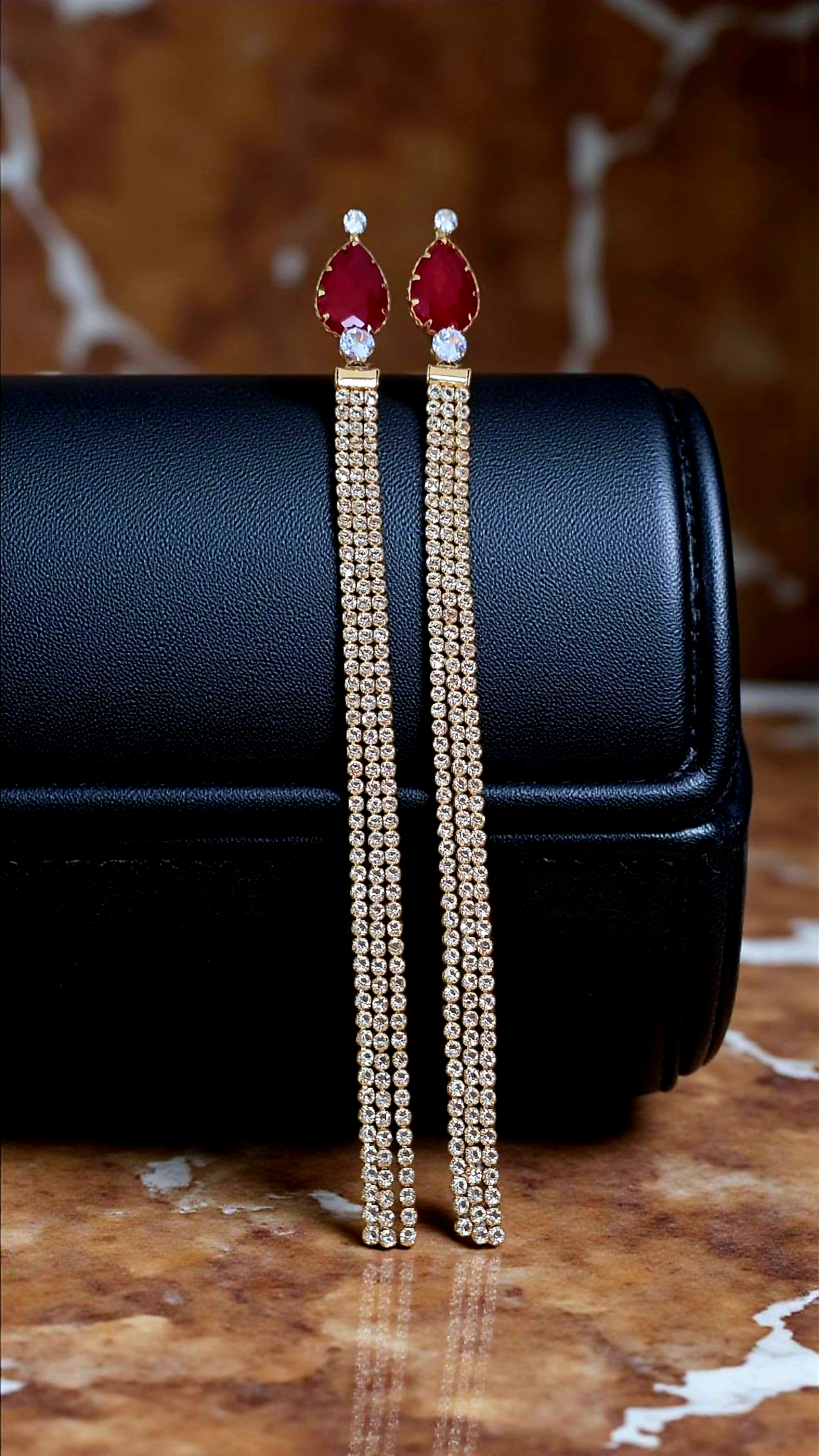 Long gold earrings with red gemstones on a black box with a marble background