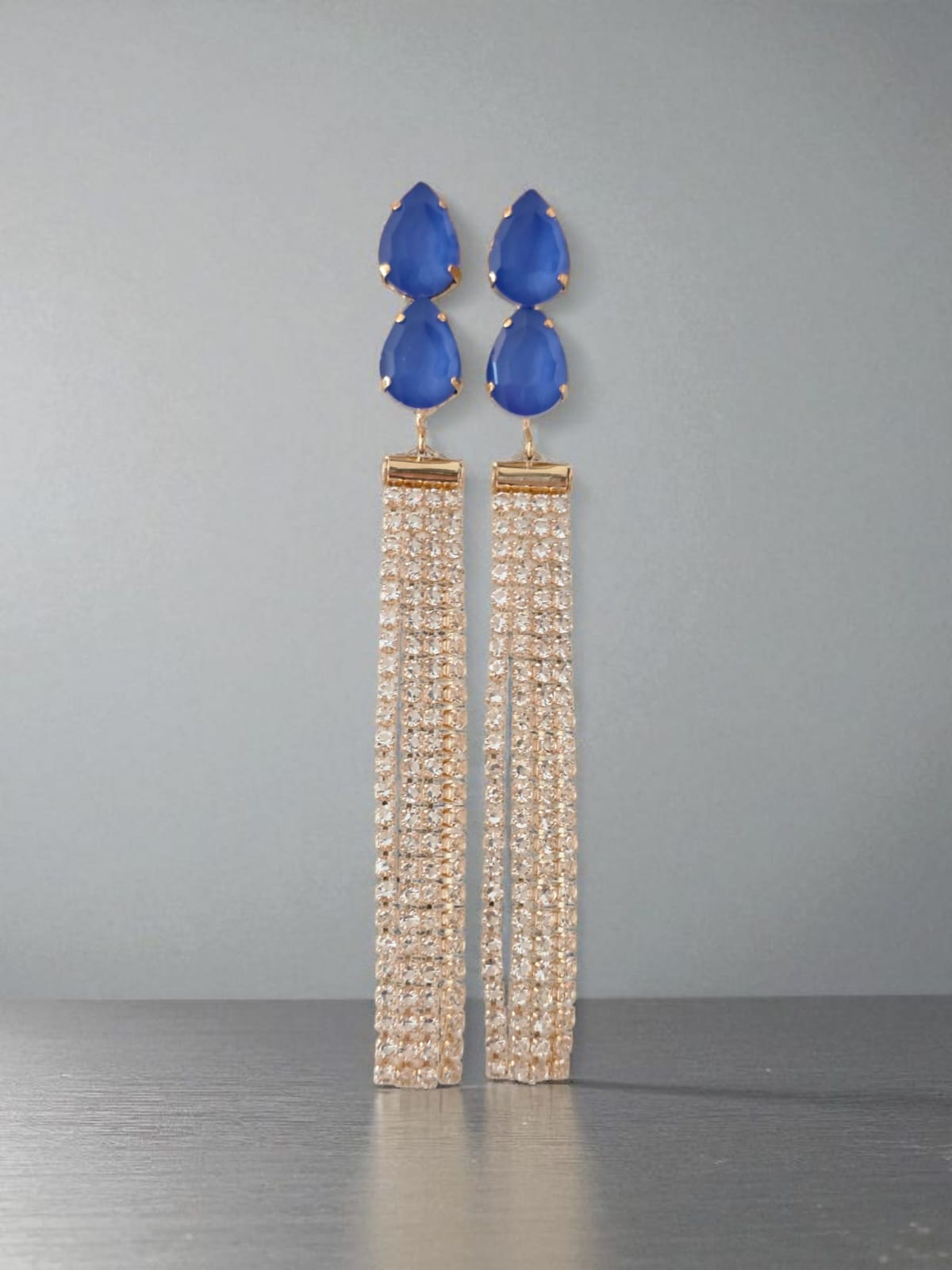 Long earrings with blue teardrop-shaped stones and gold tassels on a gray background